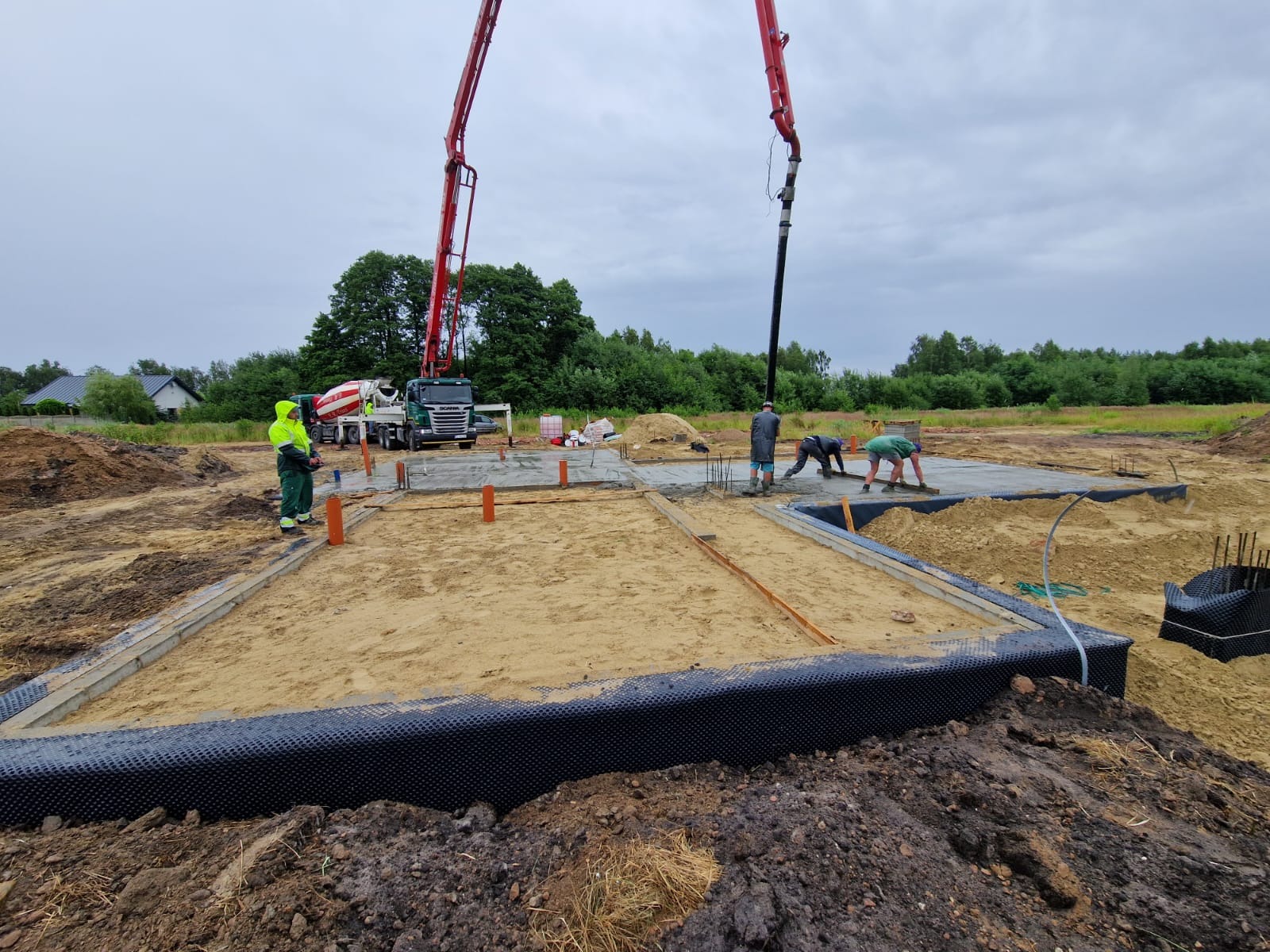 Wylewanie betonu na przygotowany fundament z izolacją, widoczny betonowóz z pompą i pracownicy wyrównujący beton.
