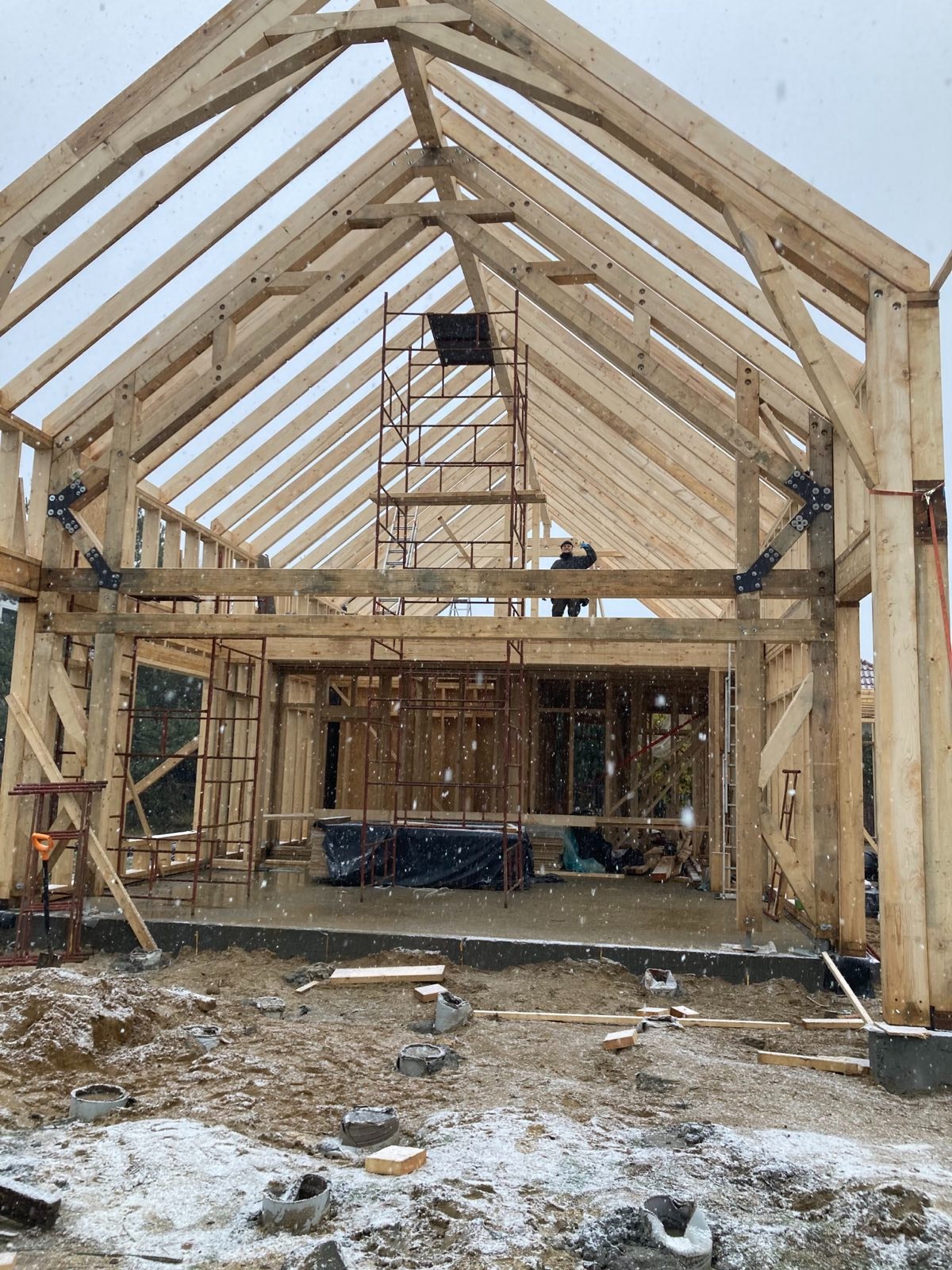 Wznoszenie drewnianego szkieletu domu w zimowej scenerii; widoczny pracownik na rusztowaniu i konstrukcja dachu w trakcie montażu.