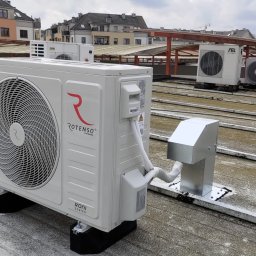 Tomasz Picho E-Klimatyzacje - Biała jednostka zewnętrzna klimatyzacji marki Rotenso, model Roni Series, zamontowana na płaskim dachu z widocznymi innymi jednostkami w tle i budynkami mieszkalnymi.