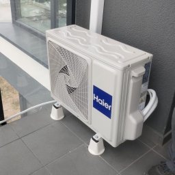 Tomasz Picho E-Klimatyzacje - Zewnętrzna jednostka klimatyzacji Haier zamontowana na balkonie, widoczne białe wsporniki i szare płytki balkonowe.