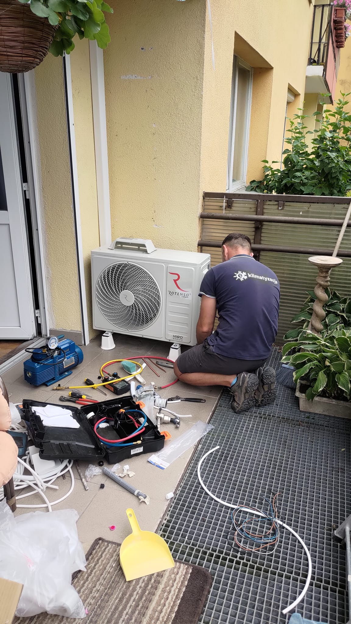 Monoblok klimatyzacji Rotenso HIRO na małych nóżkach, instalowany na zewnątrz budynku, obok otwartych drzwi balkonowych. Instalator w ciemnej koszulce z logo firmy instalacyjnej klęczy obok...
