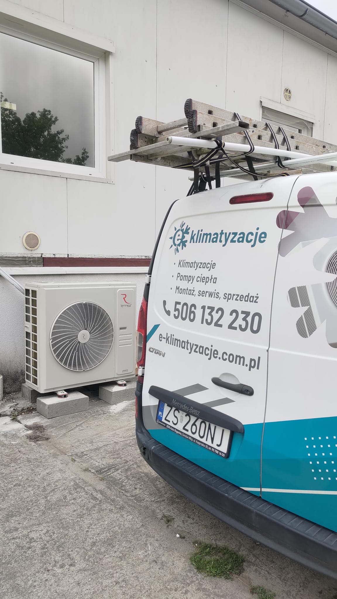 Zewnętrzna jednostka klimatyzacji Rotenso zamontowana na betonowych podstawach przy ścianie budynku, obok zaparkowany biały samochód dostawczy z reklamą firmy zajmującej się klimatyzacją i numerem...