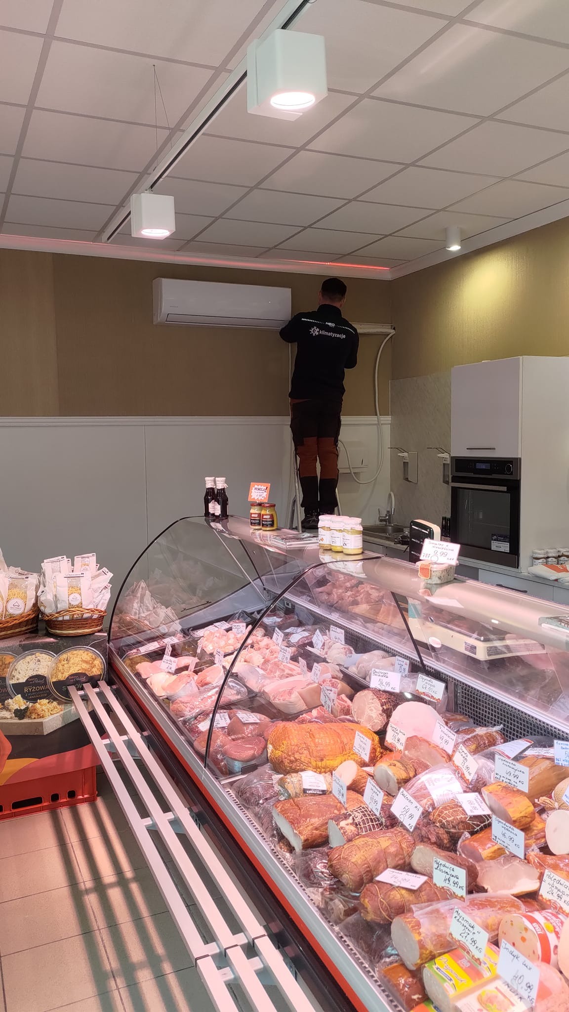 Instalator na drabinie montuje klimatyzator w sklepie spożywczym, widoczna lada chłodnicza z wędlinami i mięsem.