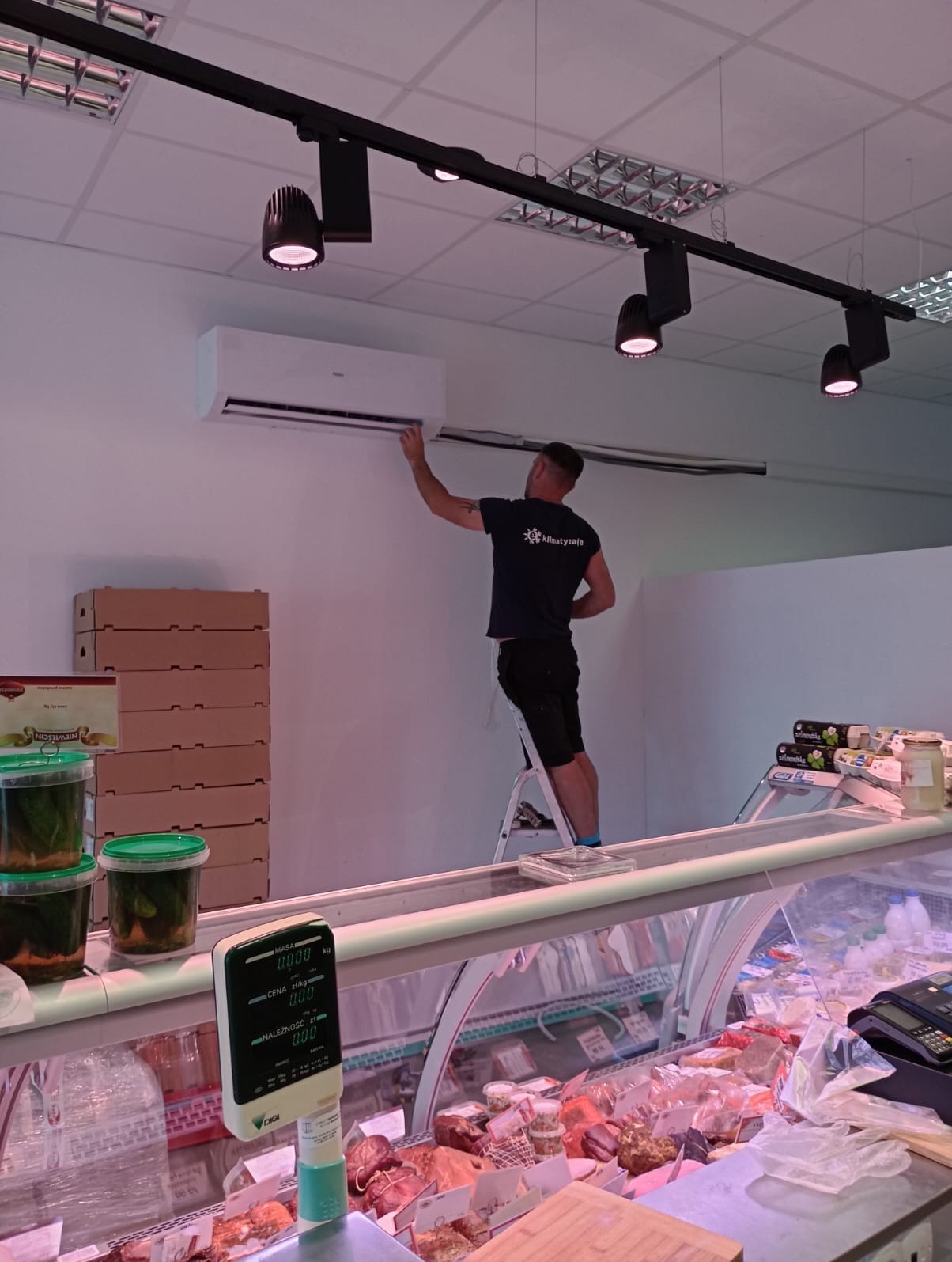 Mężczyzna na drabinie, w koszulce z logo firmy, instaluje klimatyzator Haier w sklepie spożywczym, widoczne oświetlenie szynowe i produkty w ladzie chłodniczej.