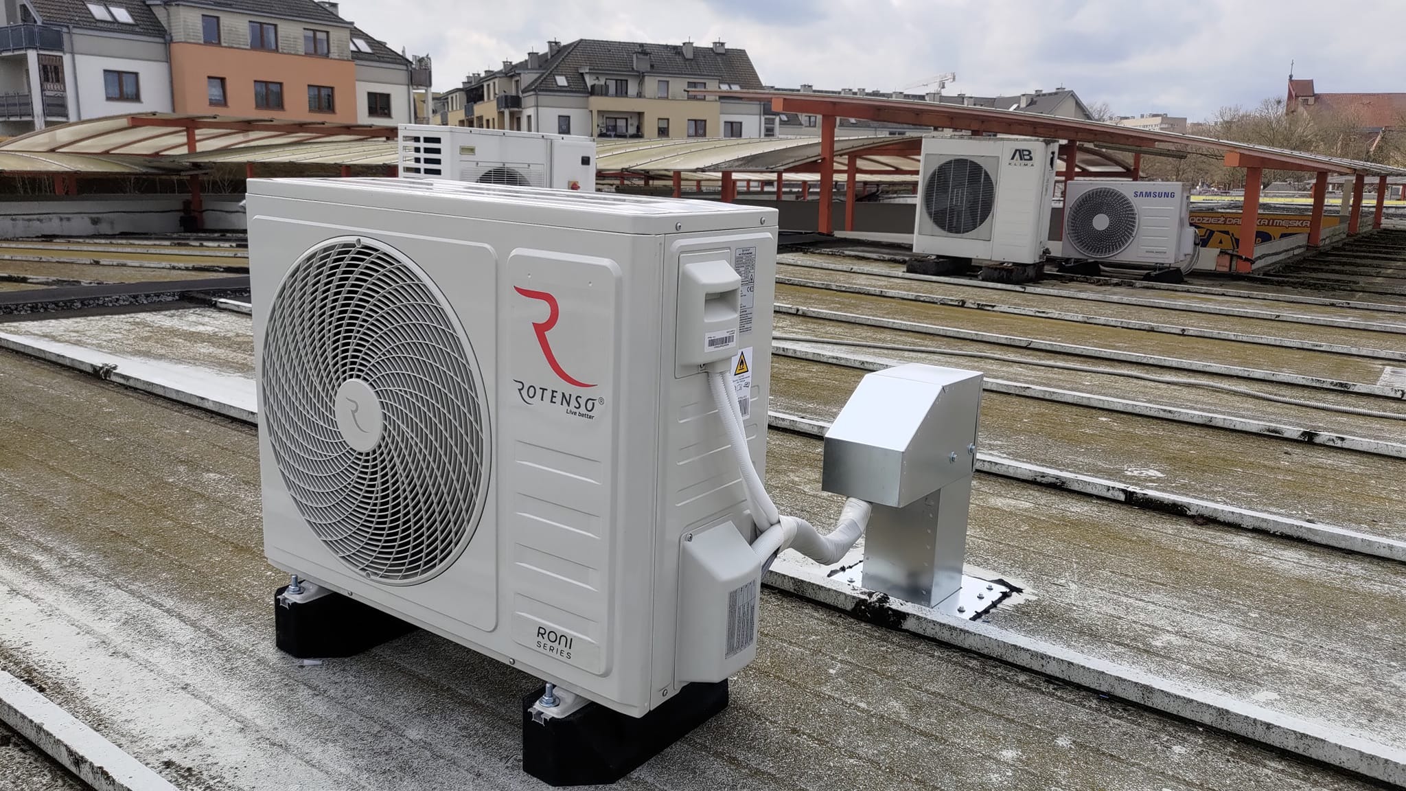 Biała jednostka zewnętrzna klimatyzacji marki Rotenso, model Roni Series, zamontowana na płaskim dachu z widocznymi innymi jednostkami w tle i budynkami mieszkalnymi.