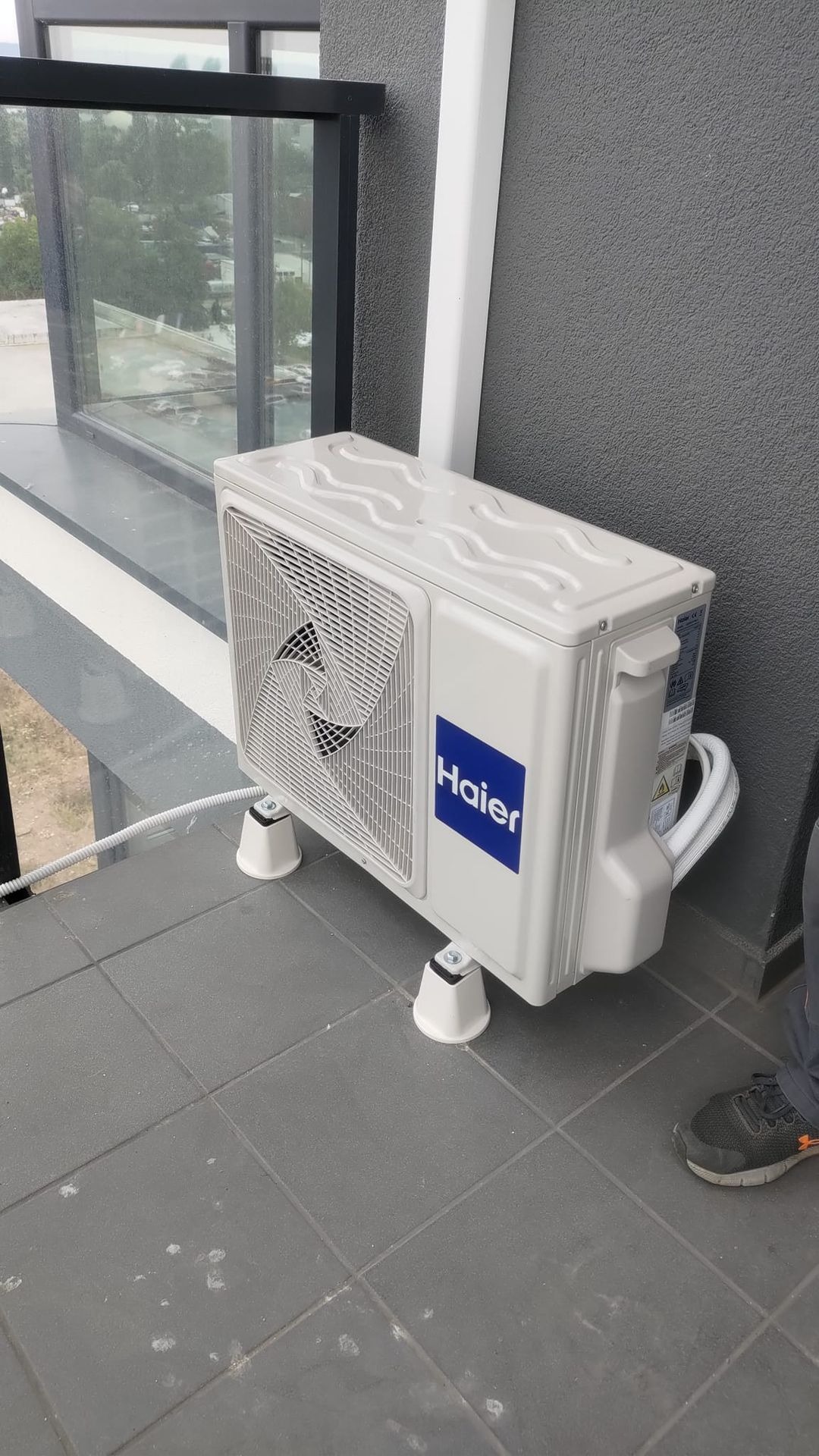 Zewnętrzna jednostka klimatyzacji Haier zamontowana na balkonie, widoczne białe wsporniki i szare płytki balkonowe.