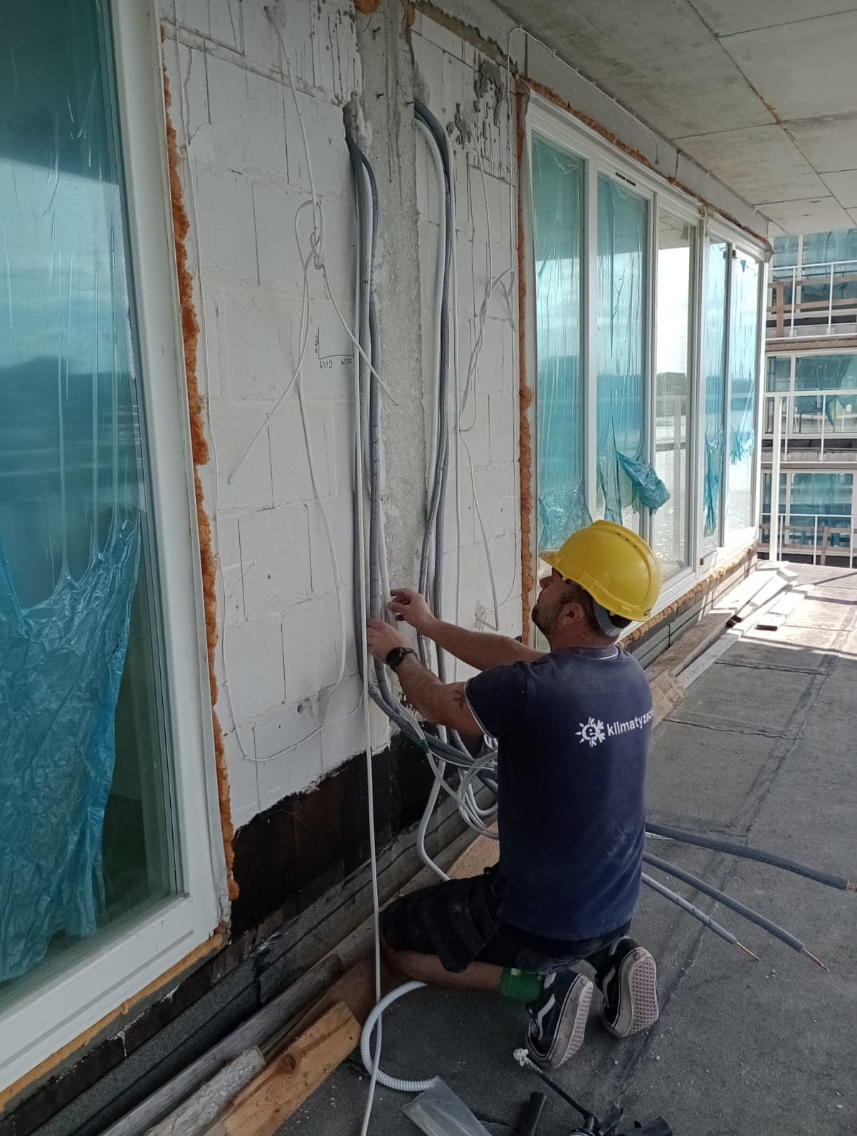 Instalator w kasku łączy przewody elektryczne na ścianie budynku z oknami, widoczne kable, pianka montażowa i logo firmy na koszulce