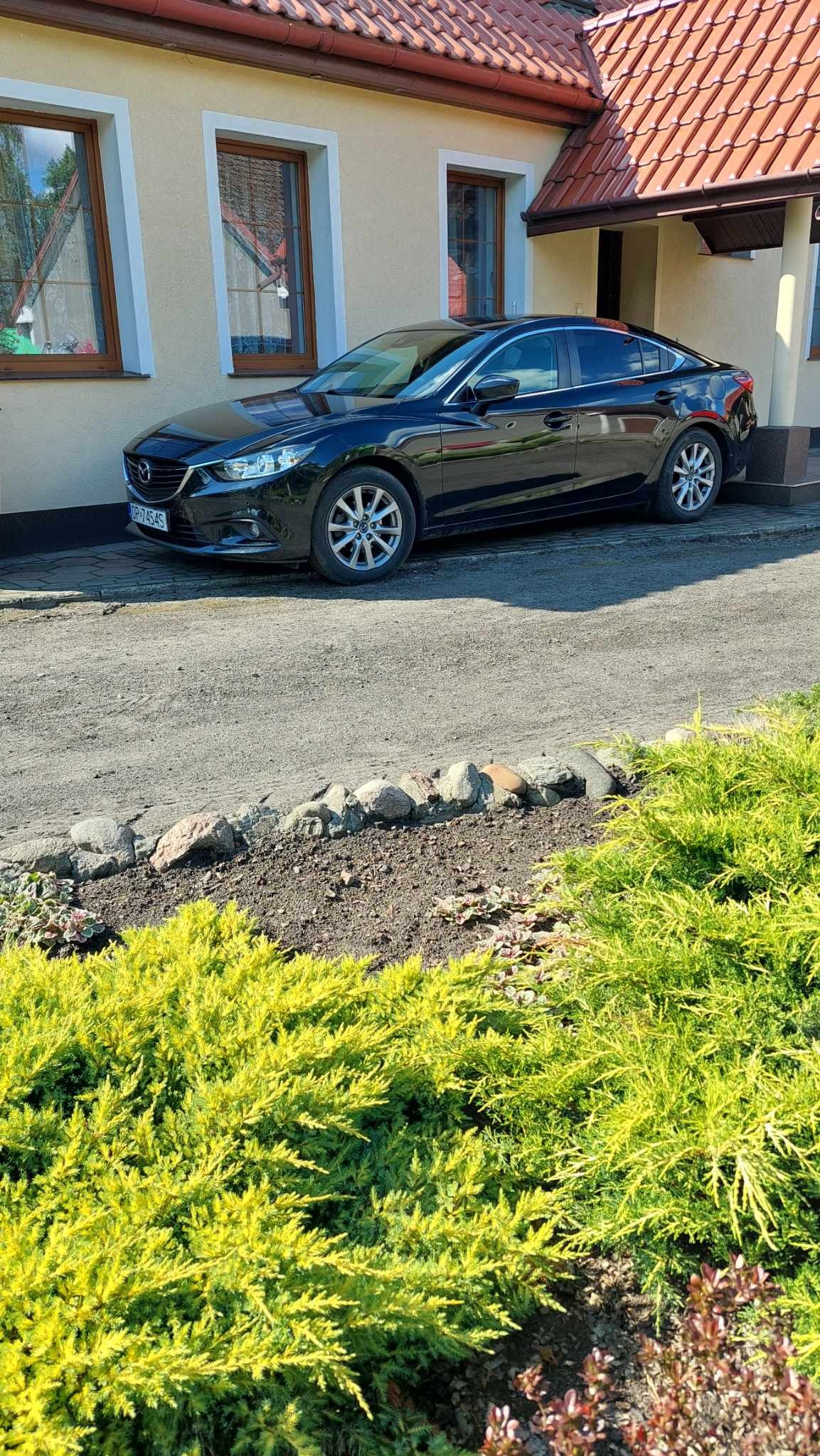 Czarny samochód Mazda zaparkowany przed beżowym domem z brązowymi oknami i czerwoną dachówką, widoczne zielone krzewy w pierwszym planie.