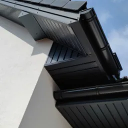 Fragment budynku z nową, ciemną elewacją, widoczne orynnowanie i podbitka dachowa w kolorze antracytowym, na tle błękitnego nieba z chmurami.