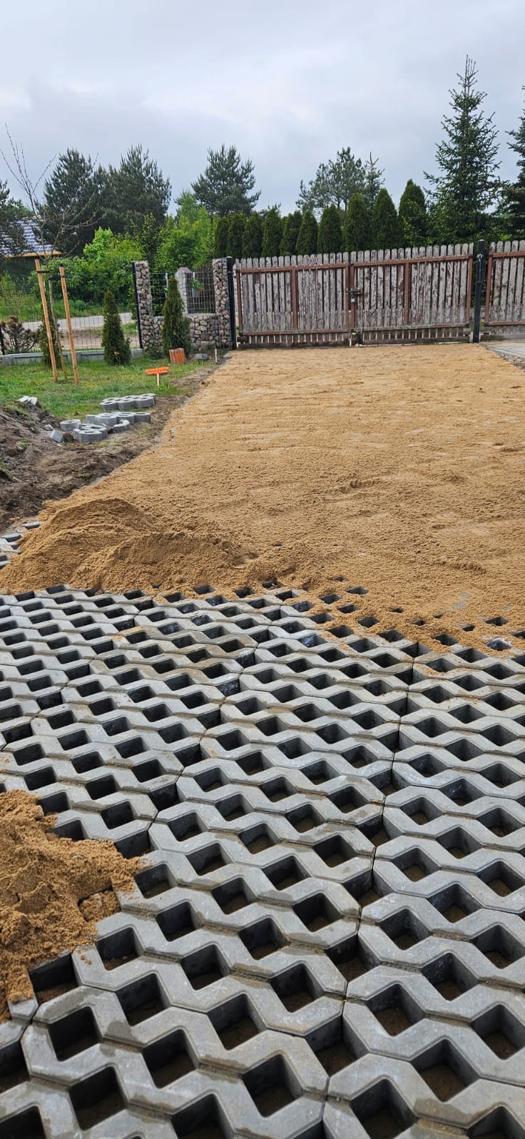 Układanie ażurowych płyt betonowych na podsypce piaskowej na podjeździe, widoczna brama w tle.
