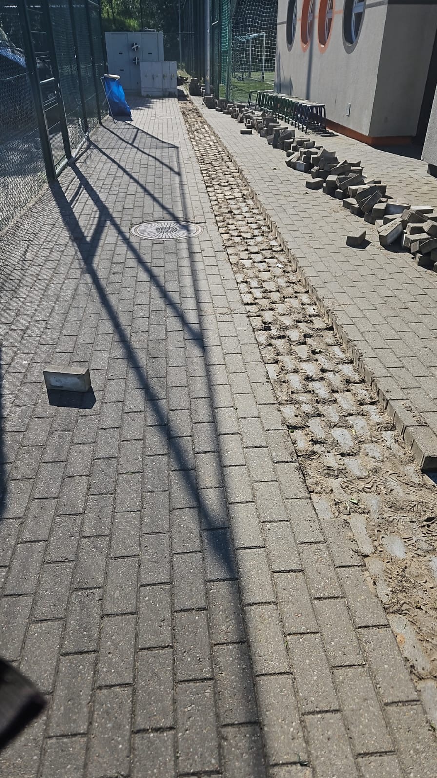 Układanie kostki brukowej na chodniku, widoczny fragment wykopu pod kostkę, stos kostki obok chodnika, cień rzucany przez siatkę ogrodzeniową.