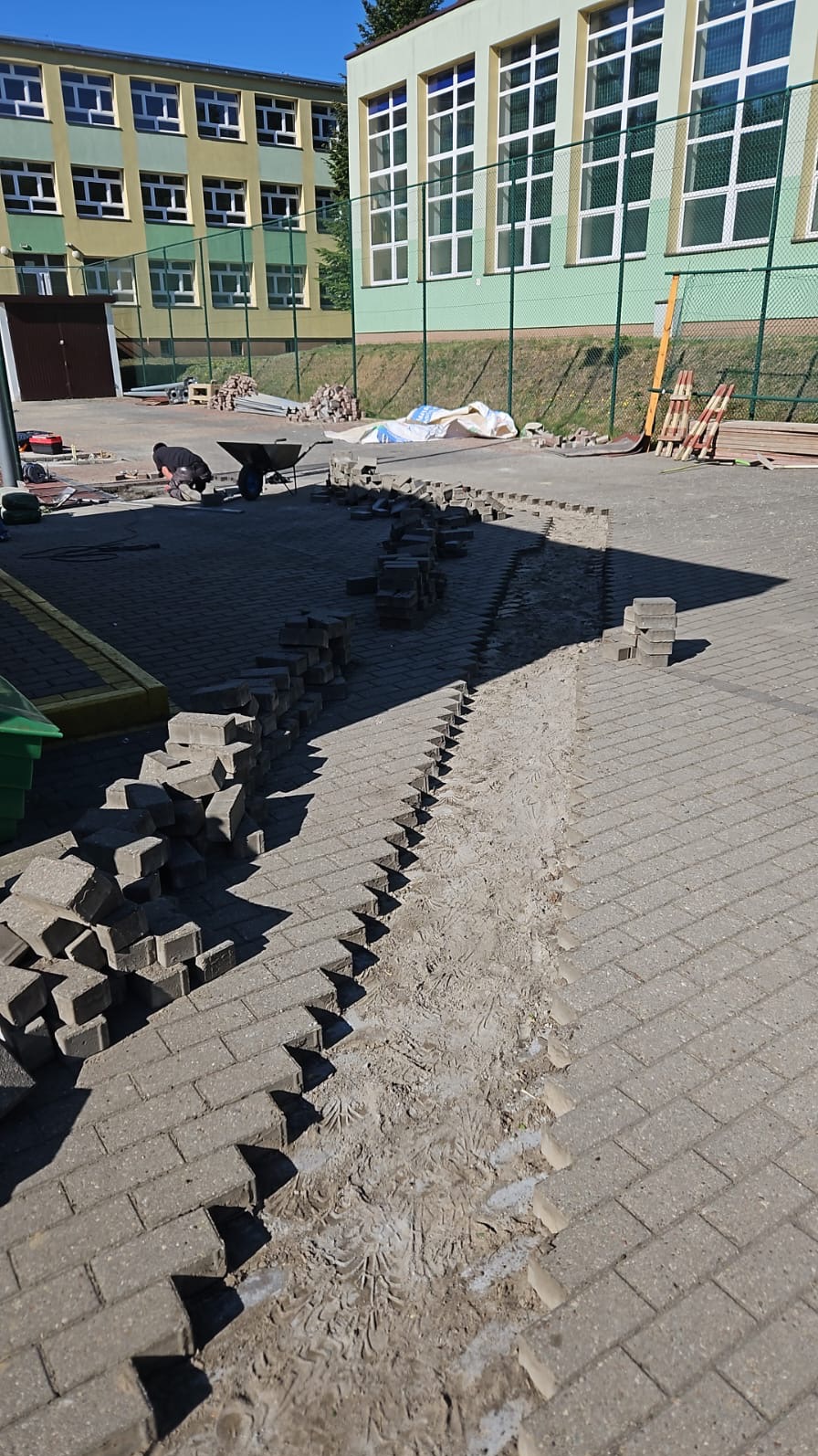 Układanie kostki brukowej na placu szkolnym, widoczny fragment wykopu z przygotowanym podłożem, ułożone rzędy kostki i stosy kostki do ułożenia, w tle budynek szkoły i pracownik z taczką.