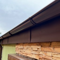 Fach Decker - Brązowe rynny i orynnowanie zamontowane na budynku z elewacją w odcieniach beżu i zieleni, widok z dołu na tle pochmurnego nieba.