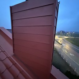 Fach Decker - Nowa, bordowa obudowa komina z poziomymi panelami na dachu z czerwonej dachówki w Inowrocławiu, widok na drogę i oświetlone miasto w tle.