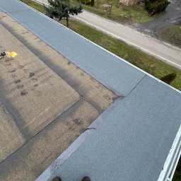 Fach Decker - Prace dekarskie na płaskim dachu z użyciem palnika, widoczne świeżo położone szare pasy papy termozgrzewalnej oraz stare, brązowe fragmenty, w tle zabudowania gospodarcze i drzewa.