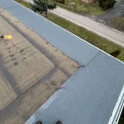 Prace dekarskie na płaskim dachu z użyciem palnika, widoczne świeżo położone szare pasy papy termozgrzewalnej oraz stare, brązowe fragmenty, w tle zabudowania gospodarcze i drzewa.