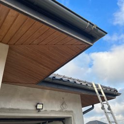 Fach Decker - Fragment domu w trakcie budowy lub remontu z widocznym fragmentem dachu z czarną rynną, drewnianą podbitką i przystawioną aluminiową drabiną na tle błękitnego nieba z chmurami.