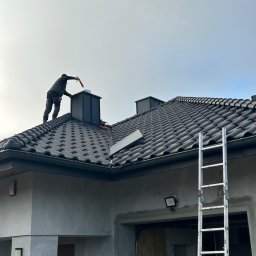 Fach Decker - Dekarz uszczelnia komin na dachu pokrytym ciemną dachówką ceramiczną, drabina oparta o rynnę, widoczny fragment elewacji domu w trakcie wykończenia.