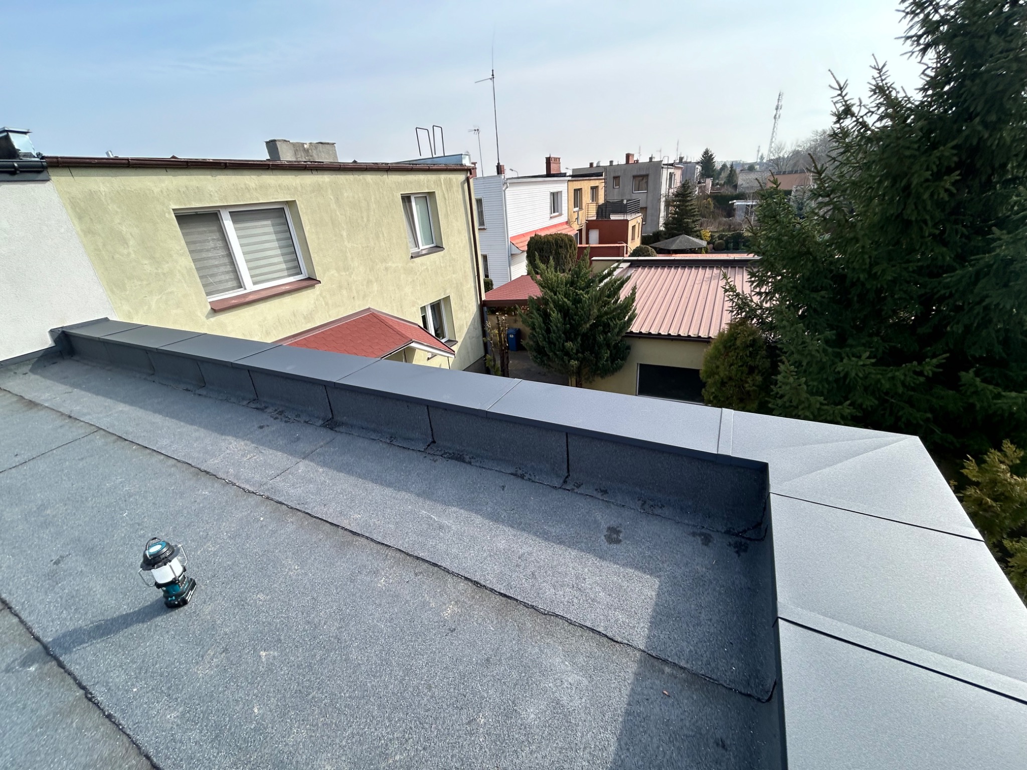 Widok z góry na płaski dach pokryty szarą papą i obróbkami blacharskimi, z latarką akumulatorową w lewym dolnym rogu, w tle zabudowa miejska i drzewa.