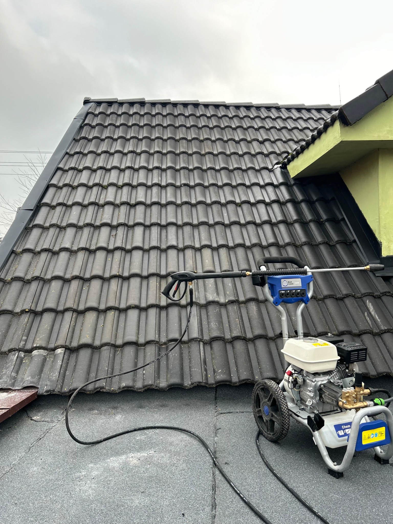 Ciemna dachówka ceramiczna czyszczona myjką ciśnieniową Blue Clean E460, widok z góry na urządzenie i fragment dachu.