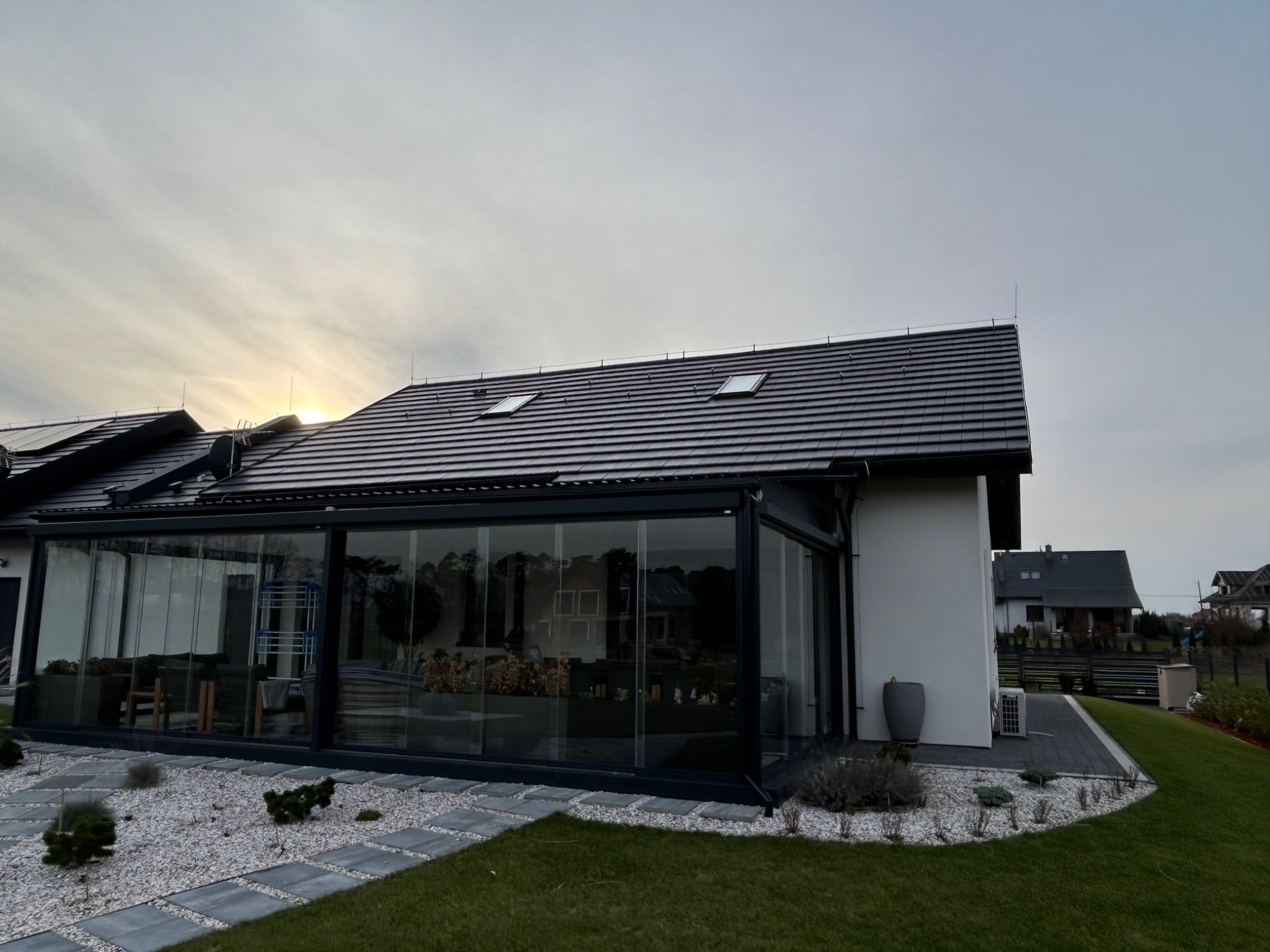 Nowoczesny dom jednorodzinny z ciemną dachówką i przeszkloną werandą, widok z ogrodu w pochmurny dzień, promienie słońca przebijają się przez chmury nad dachem.