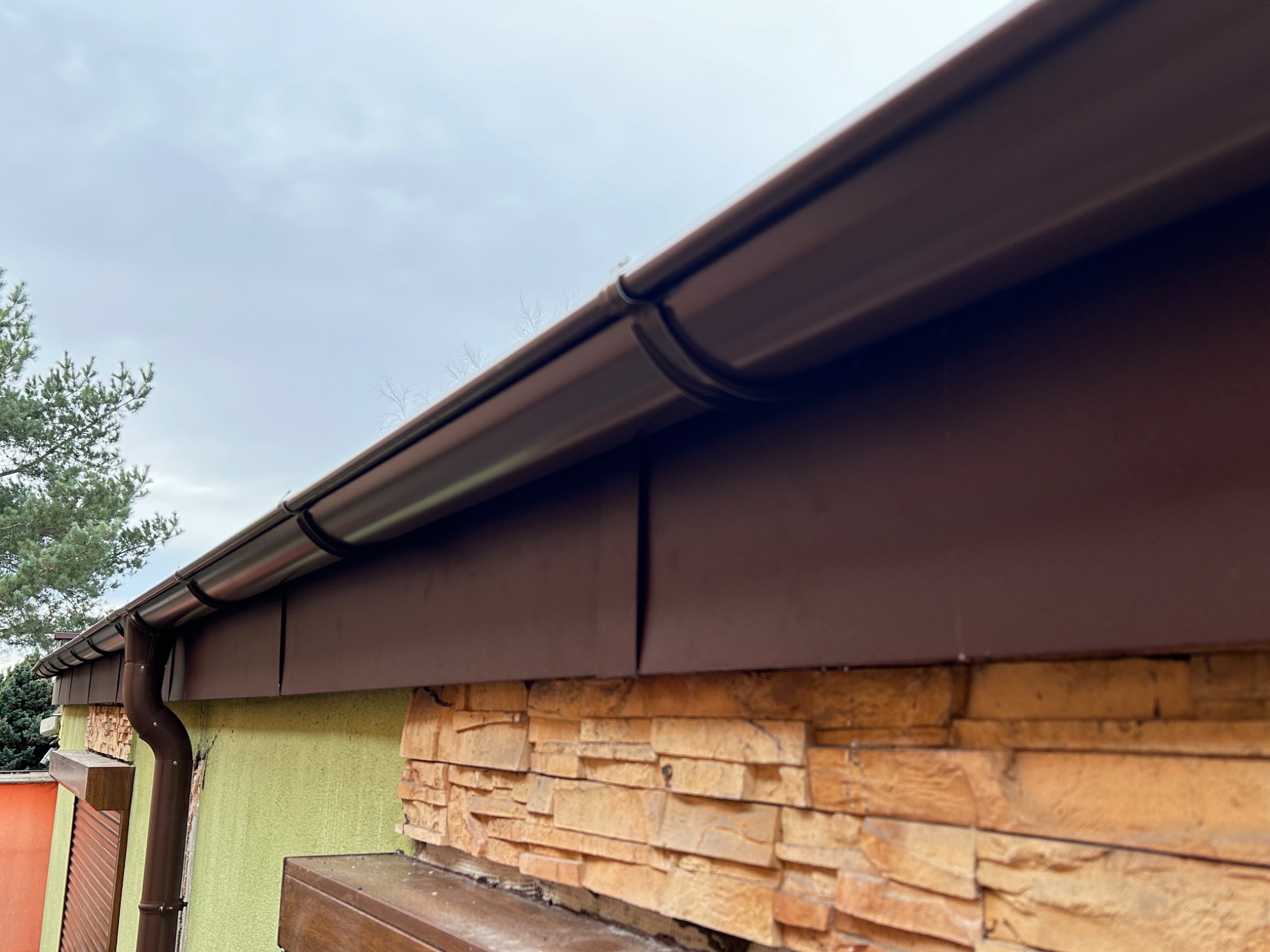 Brązowe rynny i orynnowanie zamontowane na budynku z elewacją w odcieniach beżu i zieleni, widok z dołu na tle pochmurnego nieba.
