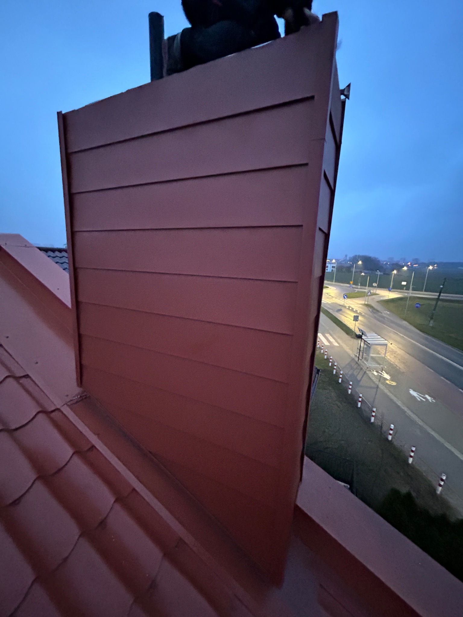 Nowa, bordowa obudowa komina z poziomymi panelami na dachu z czerwonej dachówki w Inowrocławiu, widok na drogę i oświetlone miasto w tle.