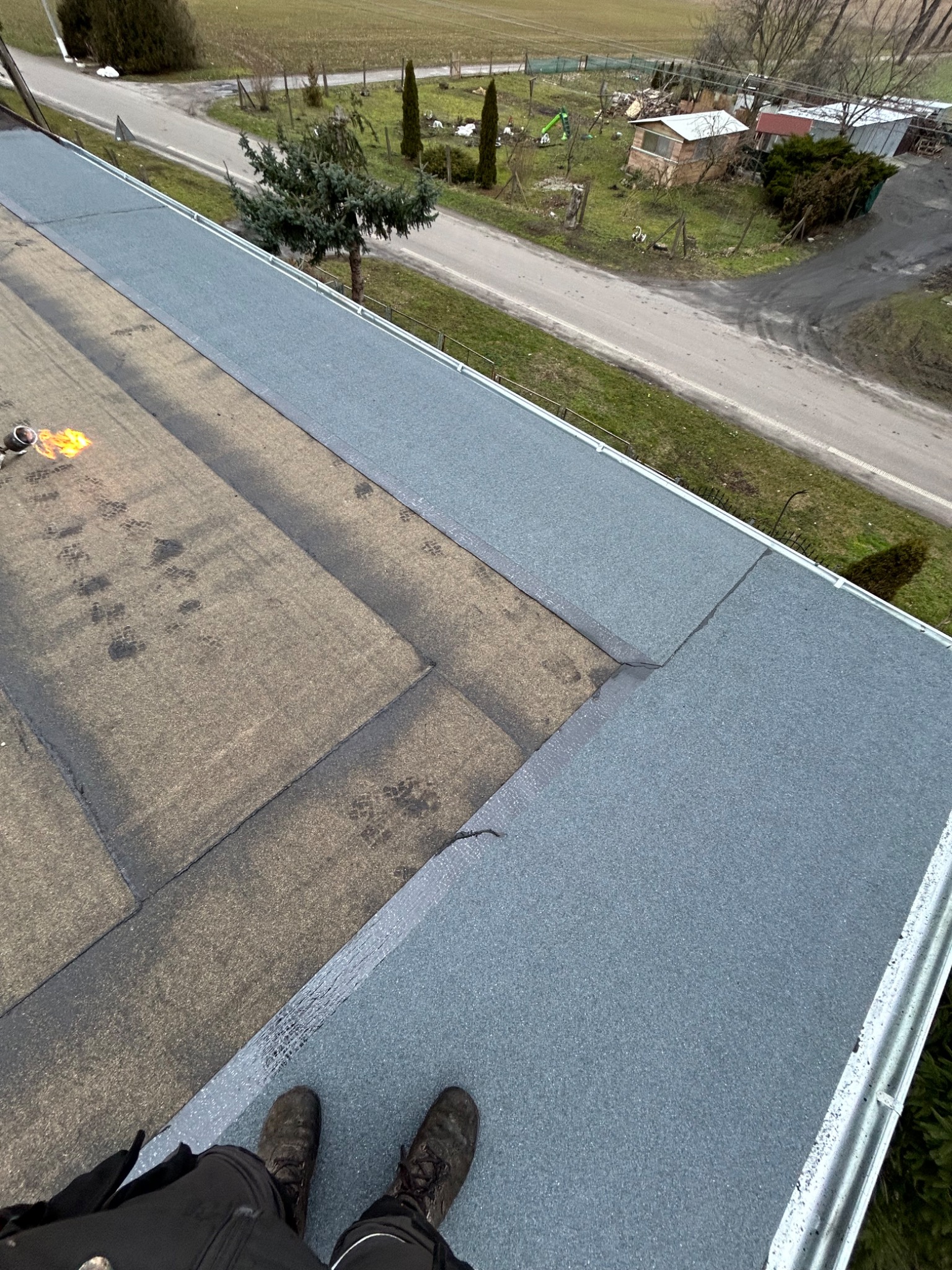 Prace dekarskie na płaskim dachu z użyciem palnika, widoczne świeżo położone szare pasy papy termozgrzewalnej oraz stare, brązowe fragmenty, w tle zabudowania gospodarcze i drzewa.