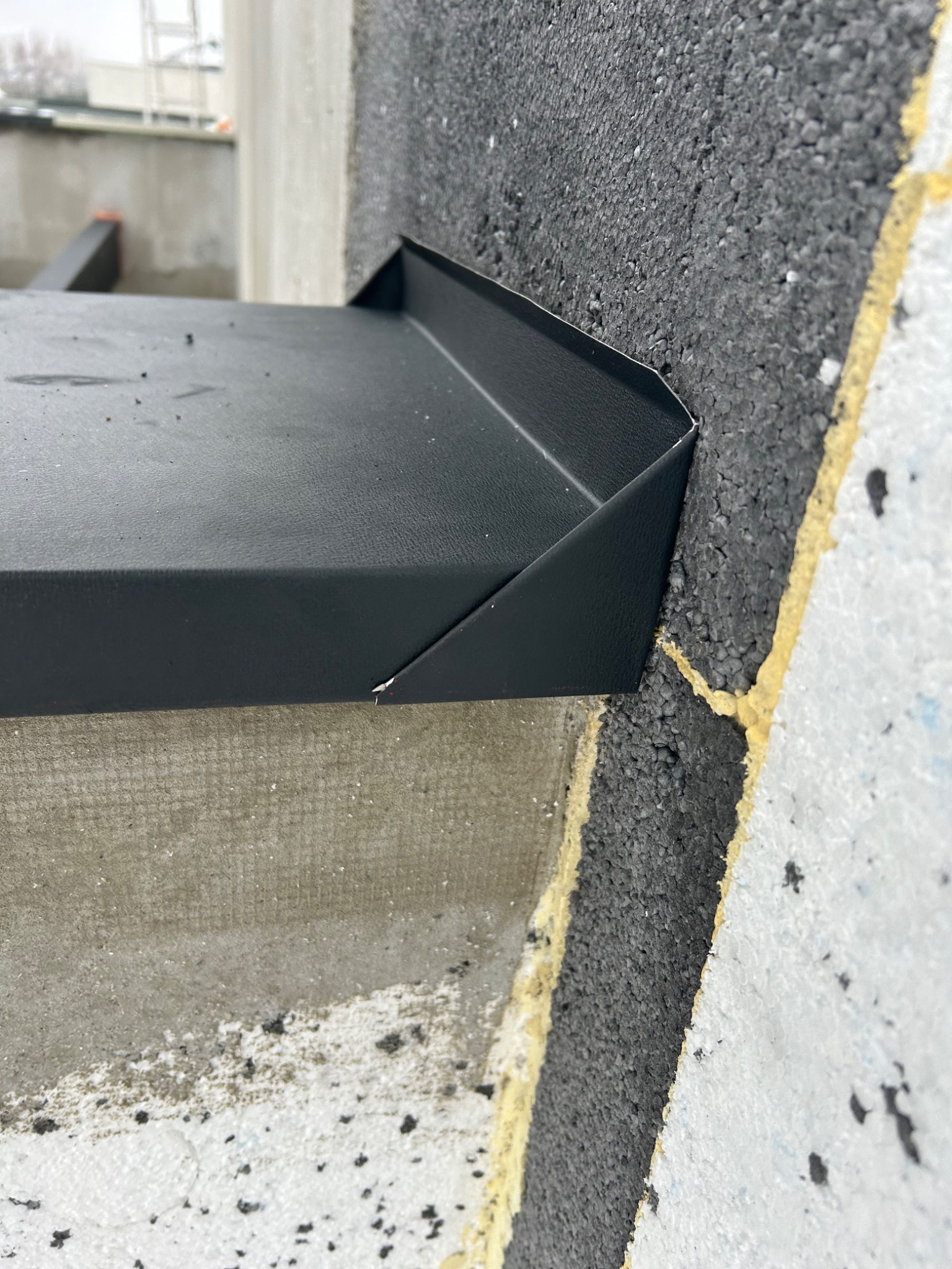 Szczegółowe ujęcie czarnej, metalowej obróbki blacharskiej, zamontowanej na styku ocieplonego budynku z elementem konstrukcyjnym, widoczne fragmenty izolacji termicznej w kolorach szarym i białym.