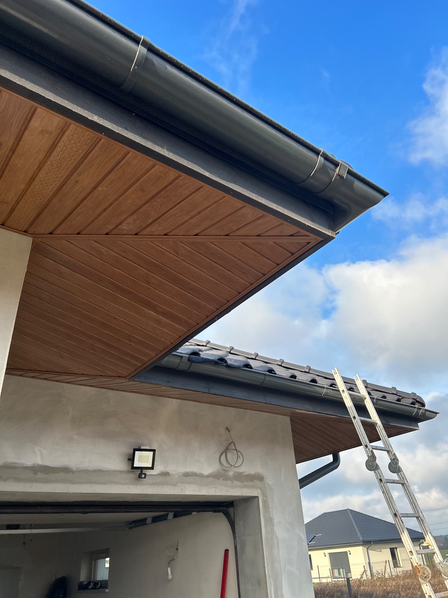 Fragment domu w trakcie budowy lub remontu z widocznym fragmentem dachu z czarną rynną, drewnianą podbitką i przystawioną aluminiową drabiną na tle błękitnego nieba z chmurami.
