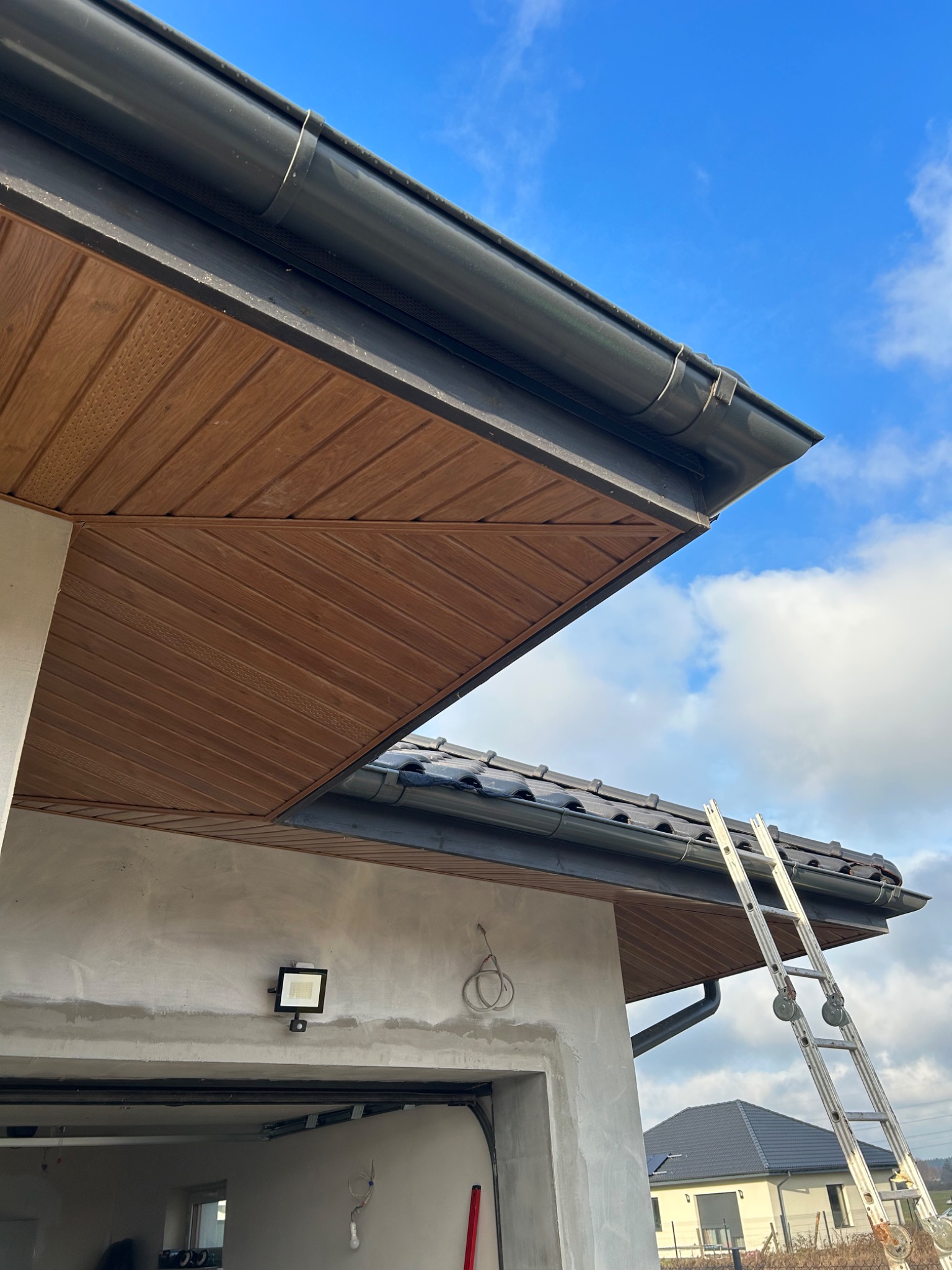 Nowe, ciemne rynny zamontowane na drewnianej podbitce dachu z użyciem drabiny na tle błękitnego nieba z chmurami.
