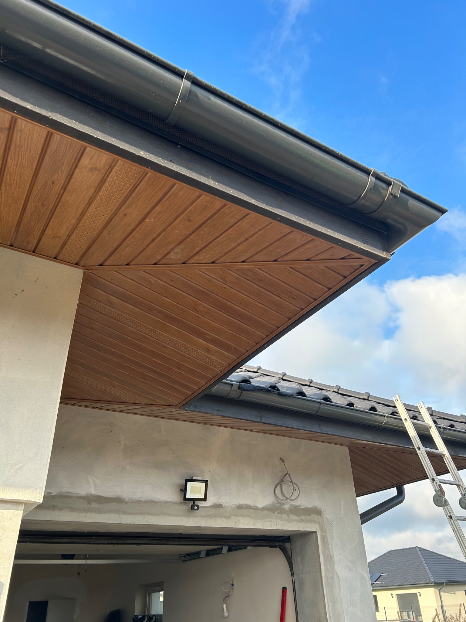 Nowe, brązowe podbitki dachowe z widocznymi czarnymi rynnami, zamontowane na budynku z szarą elewacją, drabina oparta o ścianę garażu.