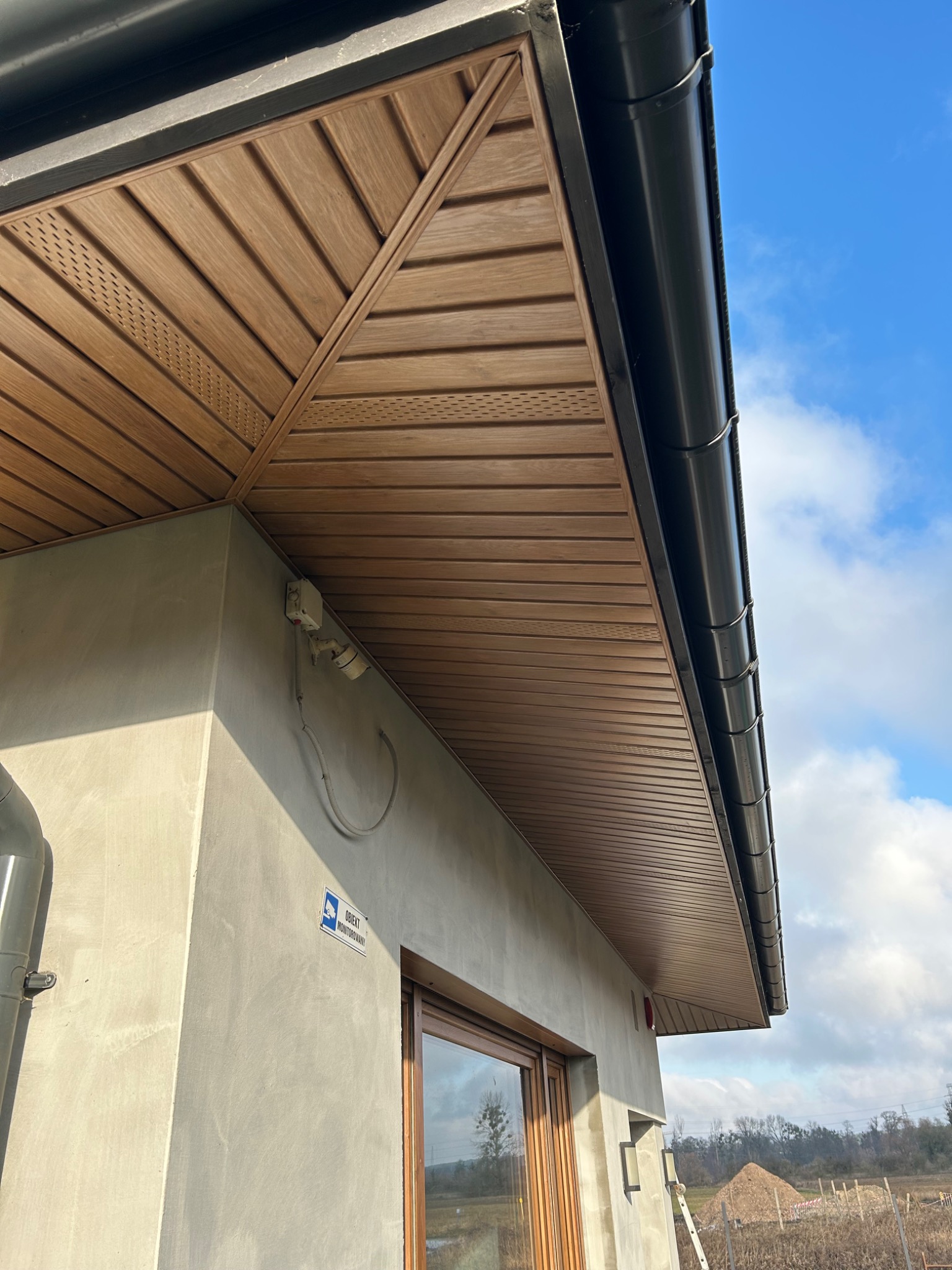 Widok z dołu na okap z brązową, panelową podbitką dachową z perforowanymi sekcjami wentylacyjnymi, czarną rynną i szarą ścianą budynku w tle, uchwycony w słoneczny dzień.