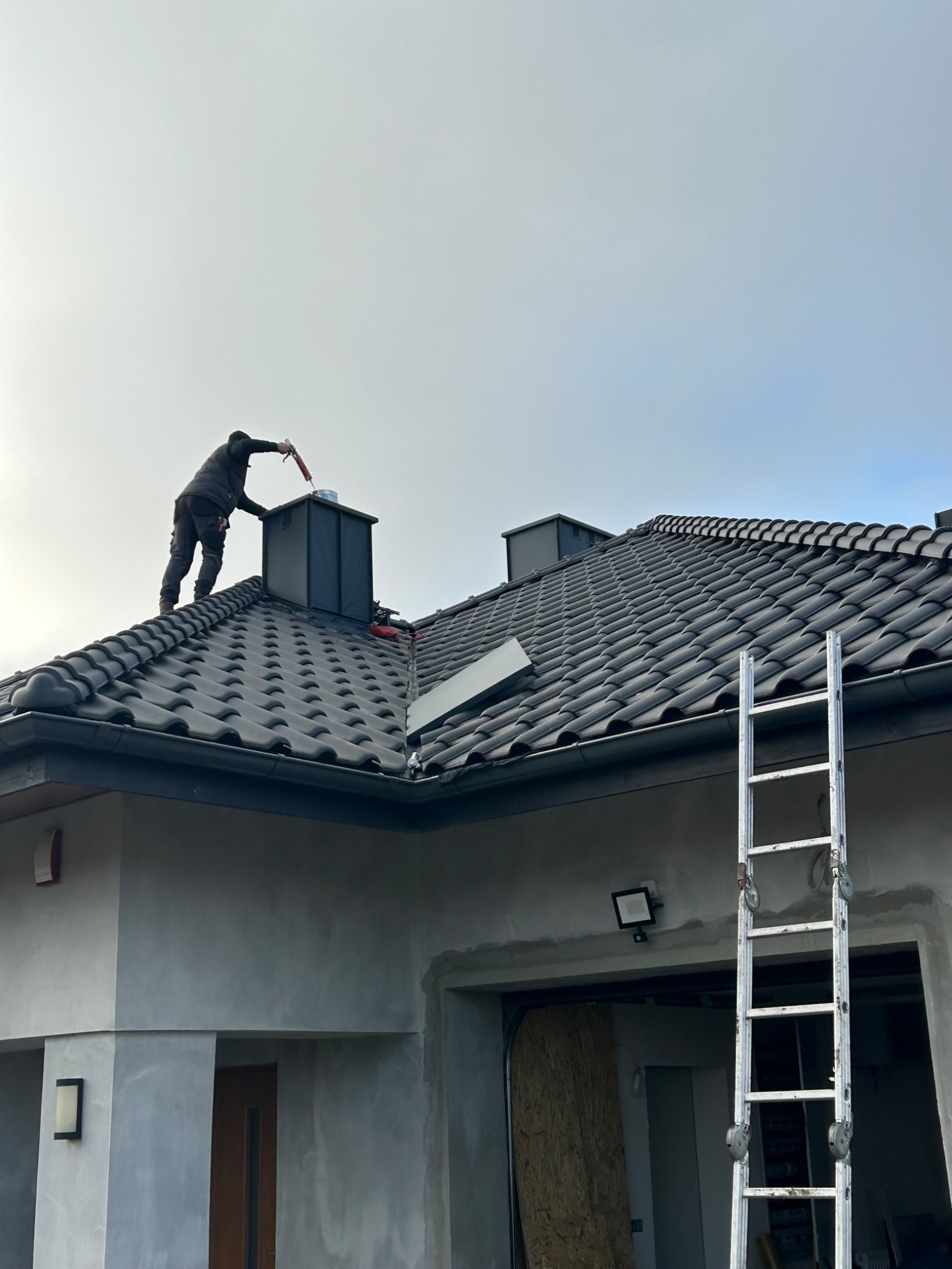 Dekarz uszczelnia komin na dachu pokrytym ciemną dachówką ceramiczną, drabina oparta o rynnę, widoczny fragment elewacji domu w trakcie wykończenia.