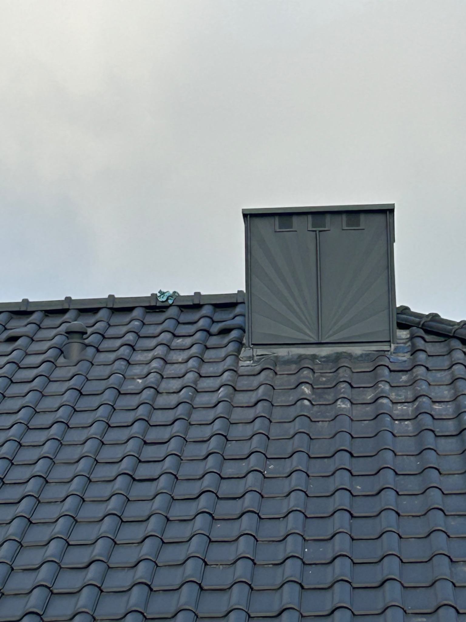 Ciemna dachówka ceramiczna z kominem wentylacyjnym z obróbką blacharską, na tle pochmurnego nieba.