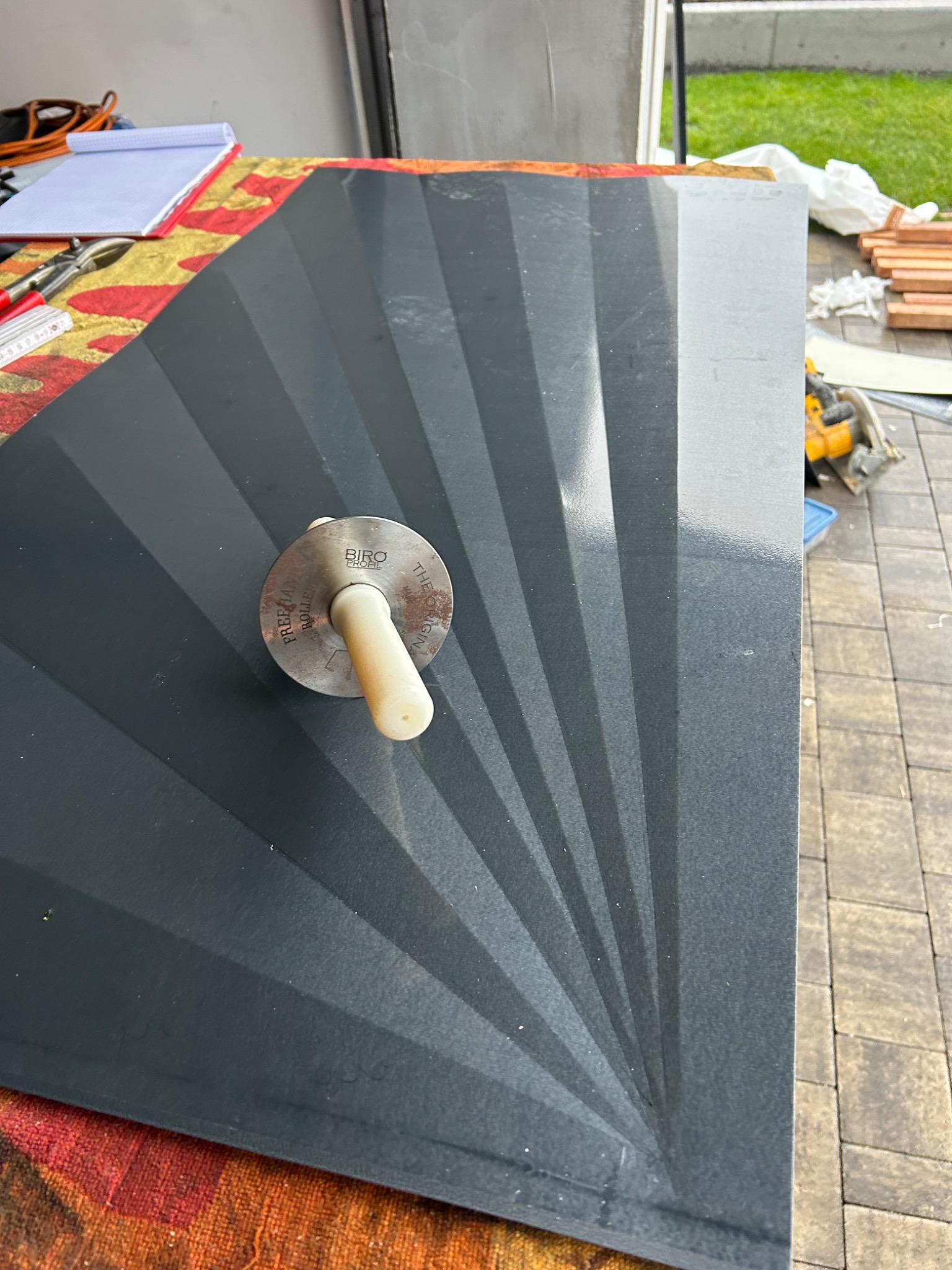 Szary panel dachowy z wytłoczonym wzorem promieni, na którym leży narzędzie dekarskie Biro Profil Freehand Roller, widoczne narzędzia i materiały budowlane w tle.