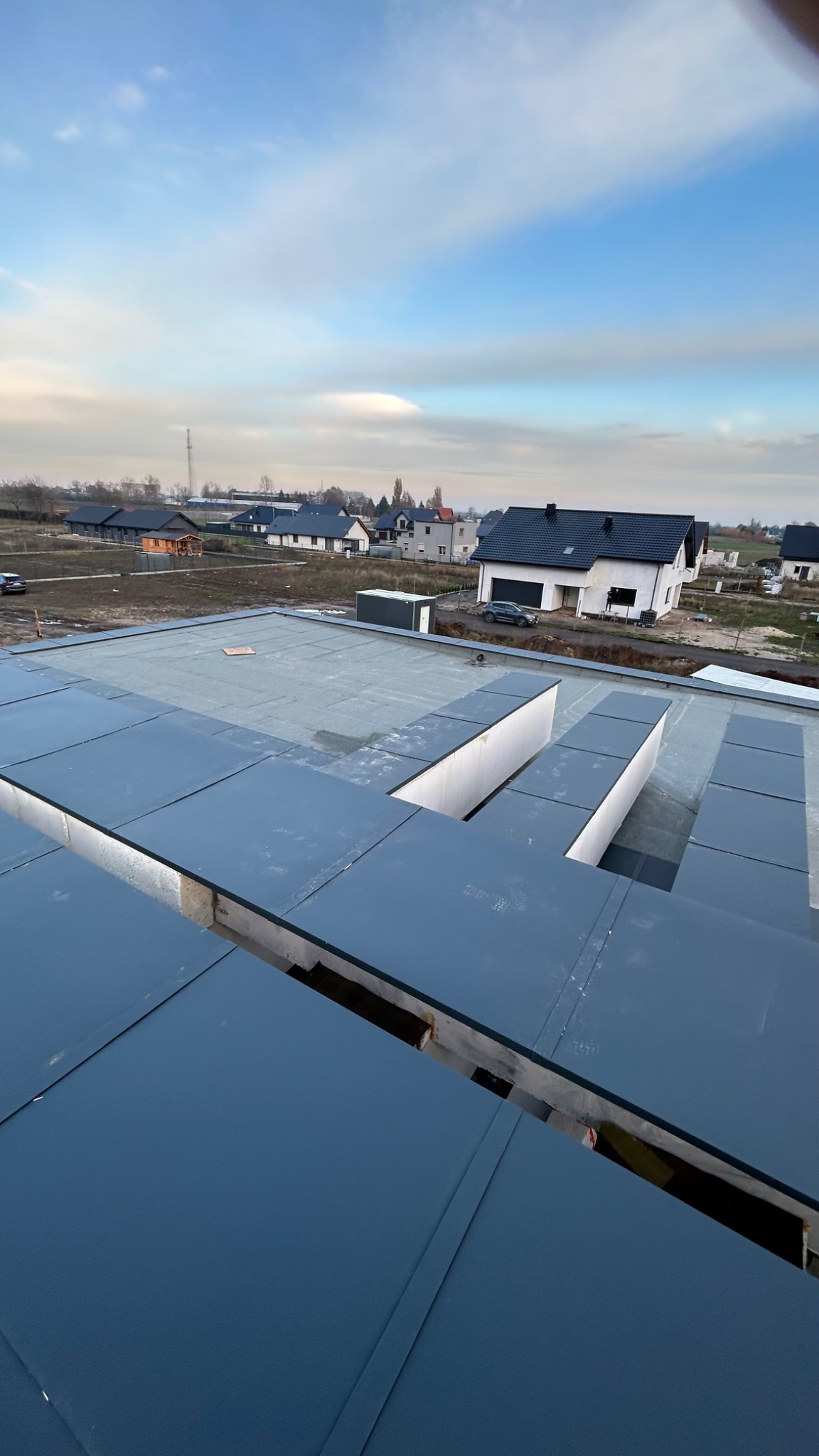 Nowoczesny, płaski dach pokryty panelami z blachy na tle zabudowy jednorodzinnej w pochmurny dzień.