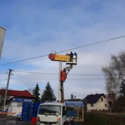 Montaż szyldu reklamowego Biedronki z użyciem podnośnika koszowego, dwóch pracowników w koszu na tle błękitnego nieba.