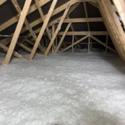 Poddasze z drewnianą konstrukcją krokwi wypełnione białą, puszystą izolacją, widoczne metalowe łączniki krokwi, ciemne poszycie dachu.