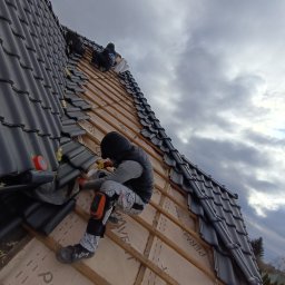 PLEPLO SPÓŁKA Z OGRANICZONĄ ODPOWIEDZIALNOŚCIĄ - Dekarze układają ciemną dachówkę na drewnianej konstrukcji dachu, widoczne membrany dachowe i pochmurne niebo w tle. Prace dekarskie w toku.