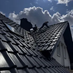 Ekipa na dachu z czarną dachówką, montuje okno dachowe na tle błękitnego nieba z chmurami. Widoczny komin i fragment elewacji budynku.