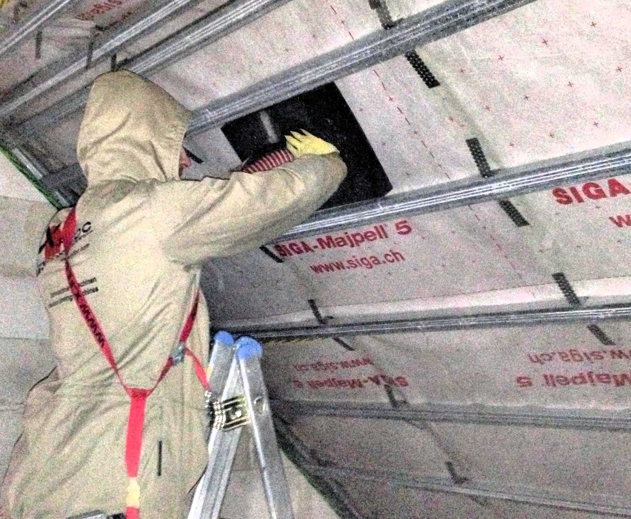 Pracownik w kombinezonie ochronnym i żółtych rękawicach, stojący na aluminiowej drabinie, montuje element w konstrukcji dachu, na folii widoczne logo Siga-Majpell.