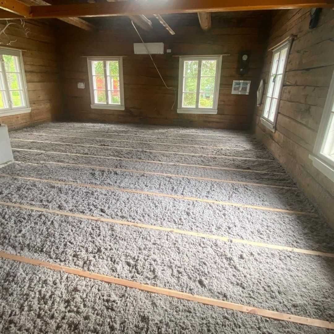 Poddasze starego drewnianego domu z widoczną warstwą szarej izolacji termicznej pomiędzy drewnianymi legarami podłogi, okna z białymi ramami w tle.