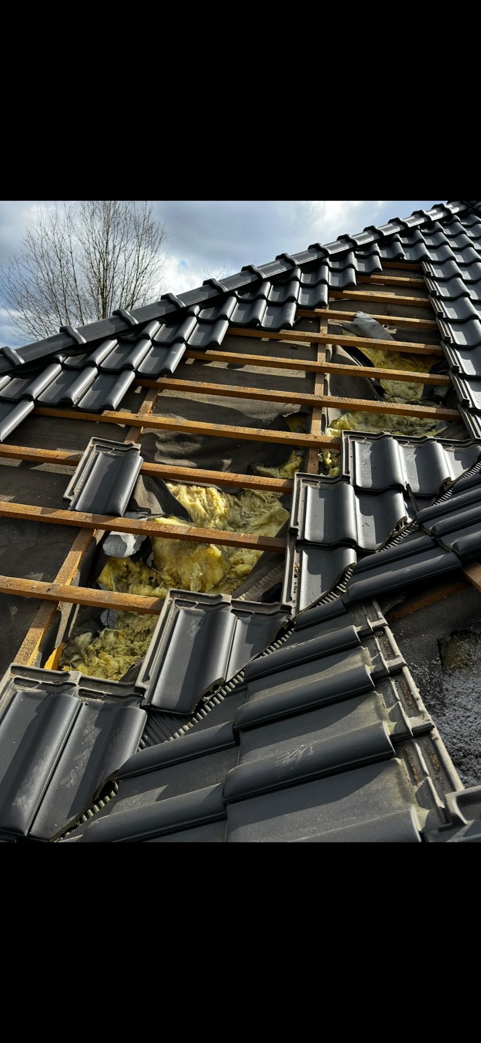 Czarny dach z widoczną izolacją z wełny mineralnej i elementami drewnianymi, częściowo pokryty dachówką. Fragment dachu w trakcie naprawy lub ocieplania.