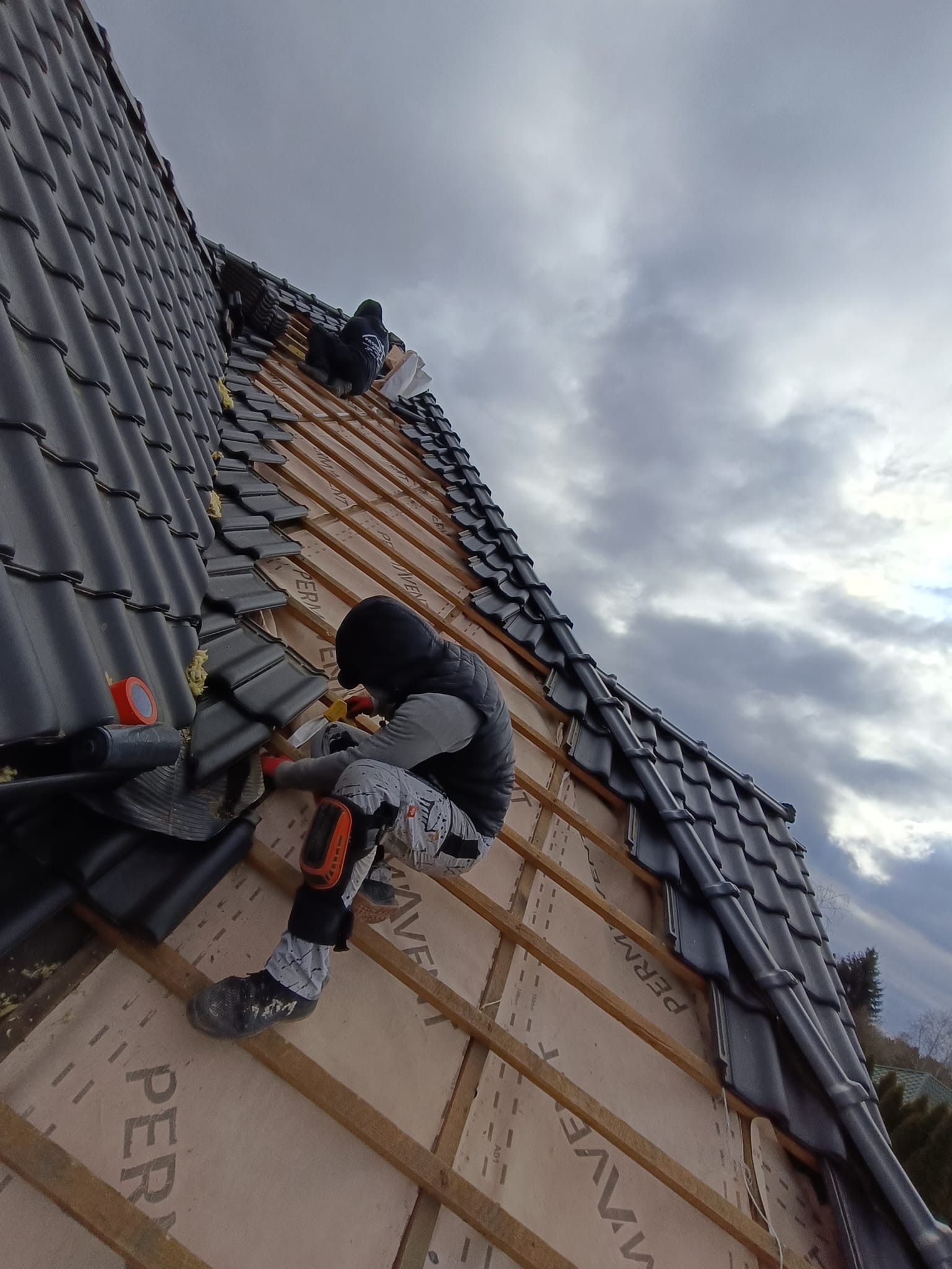 Dekarze układają ciemną dachówkę na drewnianej konstrukcji dachu, widoczne membrany dachowe i pochmurne niebo w tle. Prace dekarskie w toku.