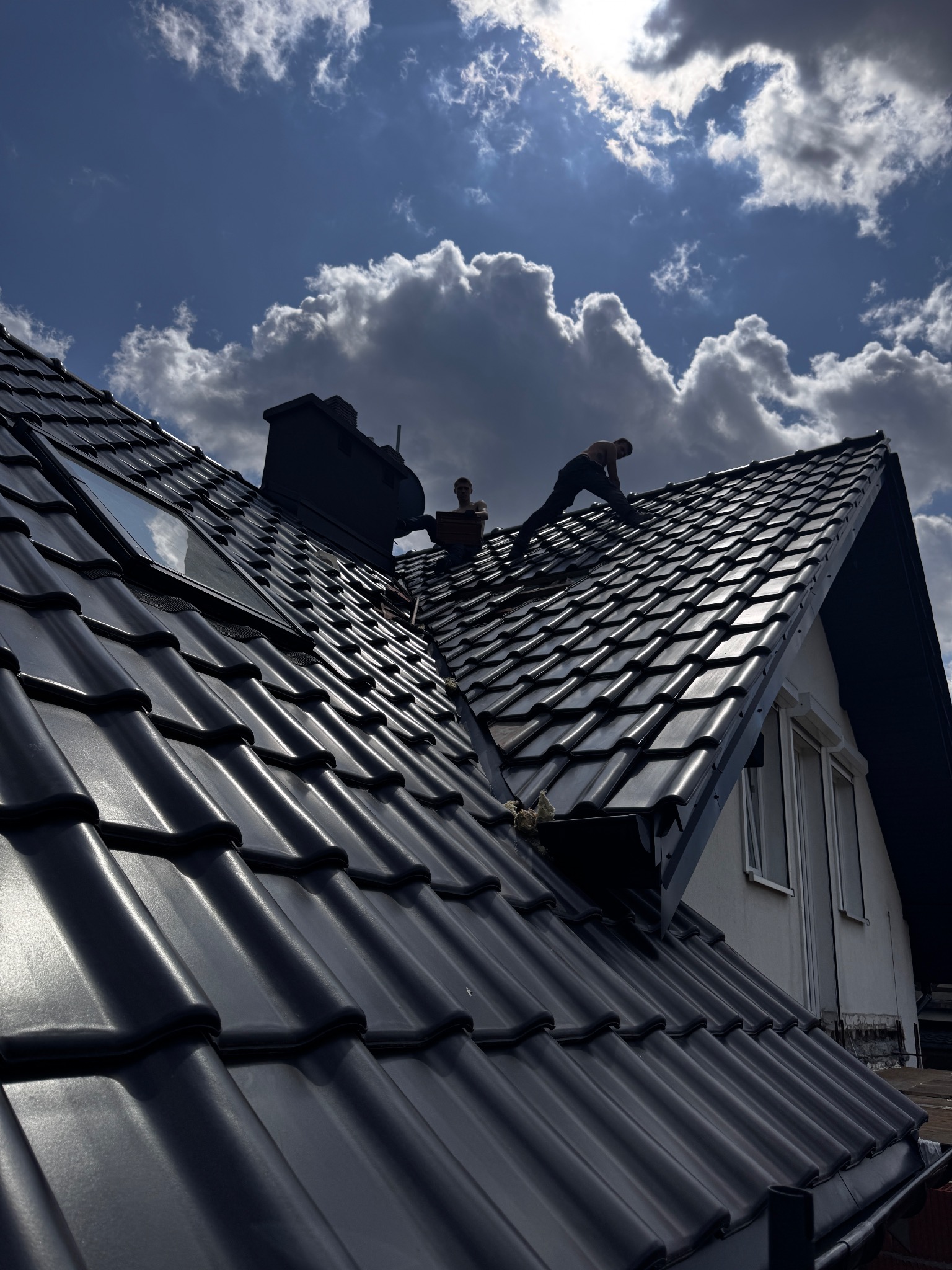 Ekipa na dachu z czarną dachówką, montuje okno dachowe na tle błękitnego nieba z chmurami. Widoczny komin i fragment elewacji budynku.