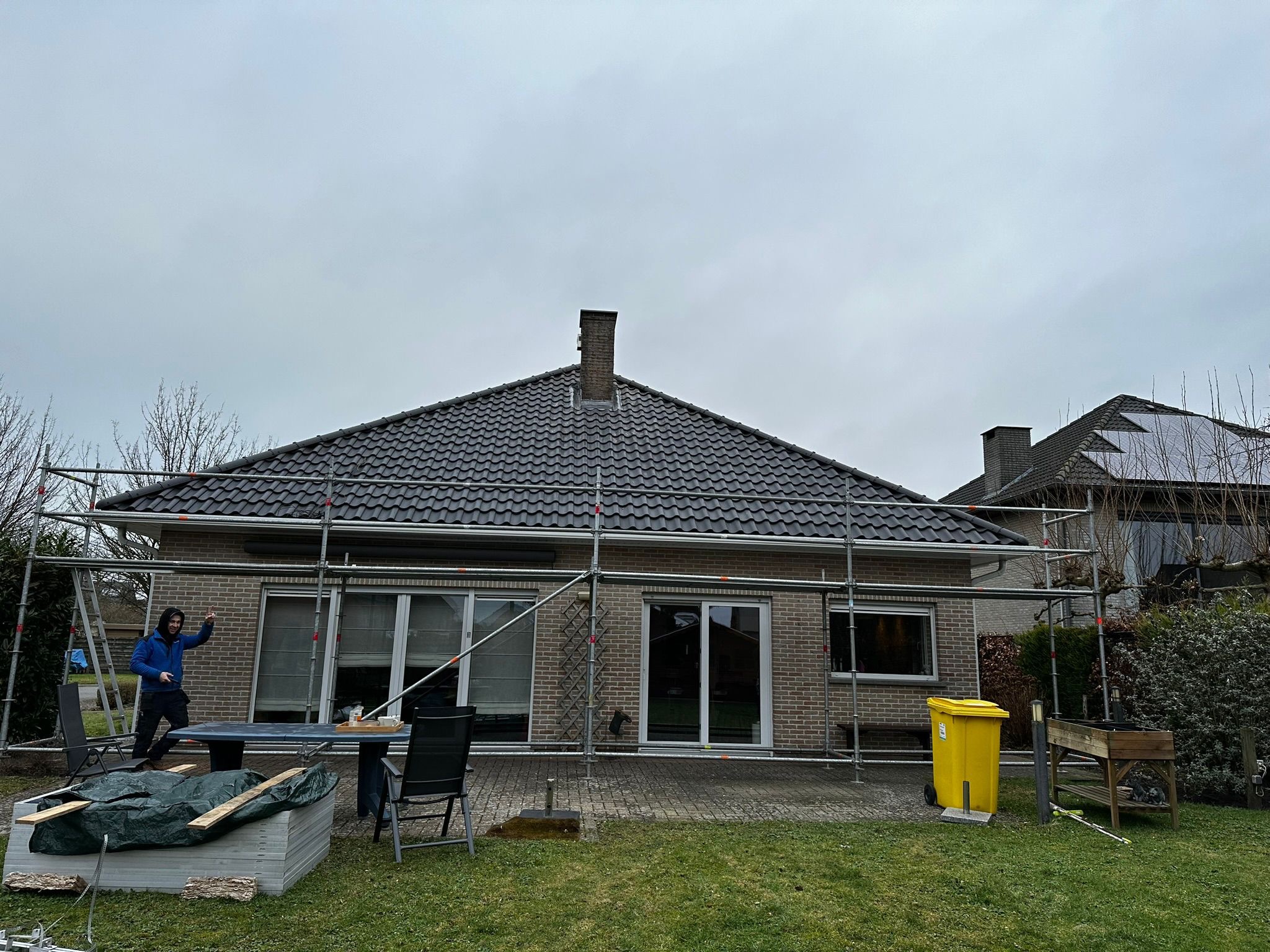 Dom jednorodzinny w trakcie prac na rusztowaniu, prawdopodobnie ociepleniowych. Widoczny pracownik, materiały budowlane oraz żółty pojemnik na odpady.