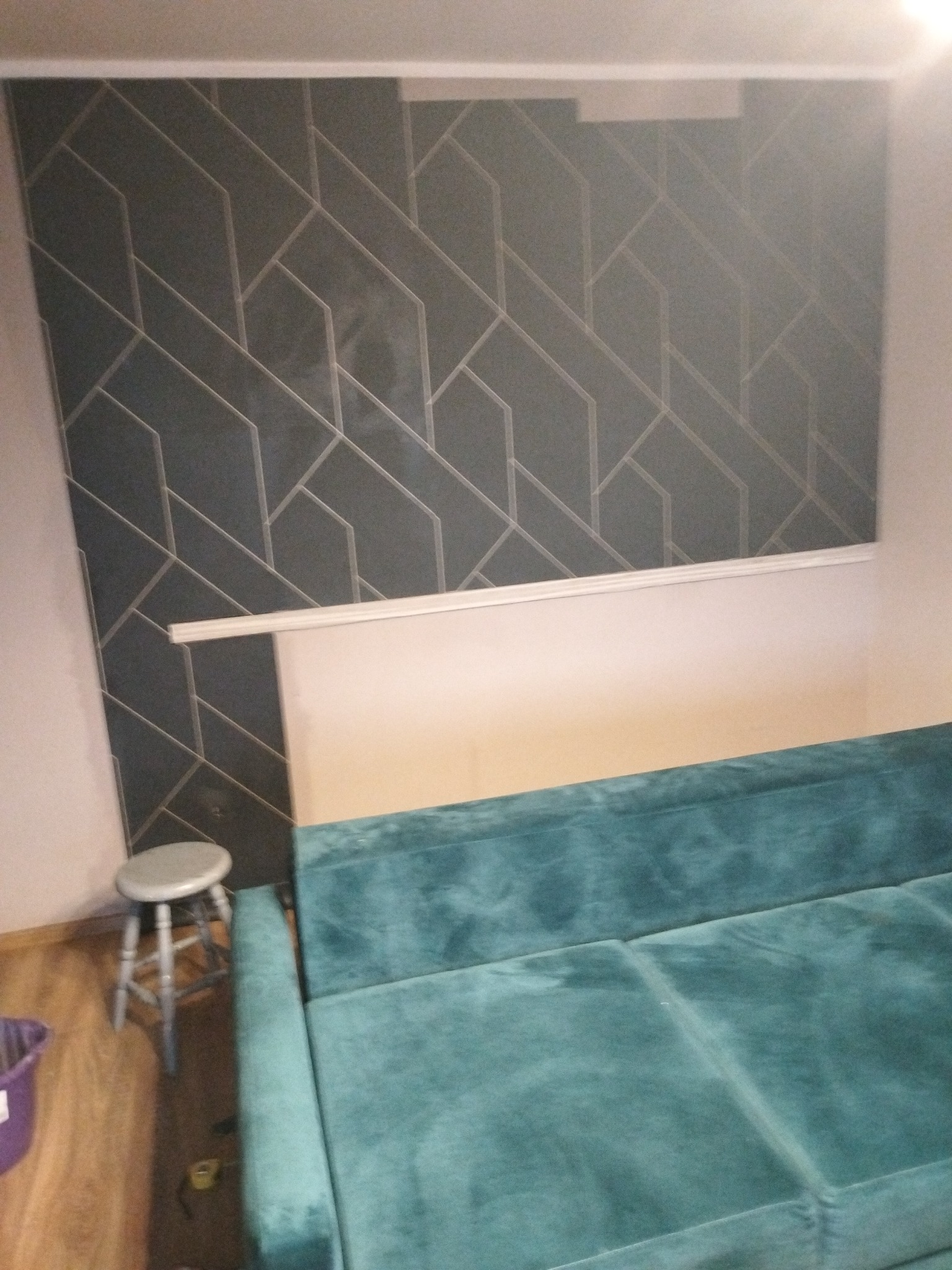 Fragment pokoju z szaro-białą tapetą w geometryczne wzory, listwą przysufitową, turkusową kanapą i drewnianym stołkiem.