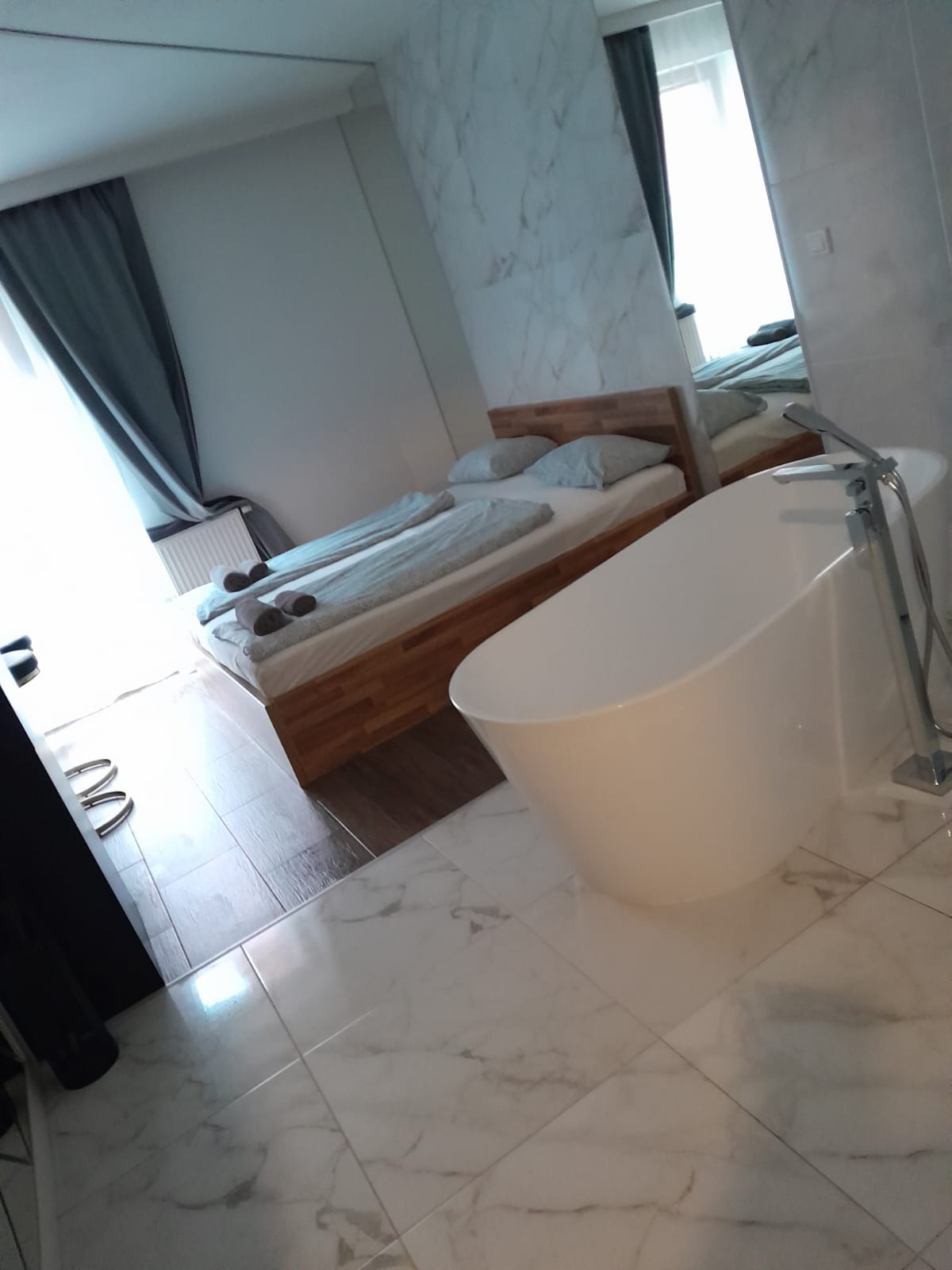 Nowoczesna łazienka z wolnostojącą wanną i marmurową okładziną ścienną, widok na sypialnię z drewnianym łóżkiem i szarymi zasłonami.