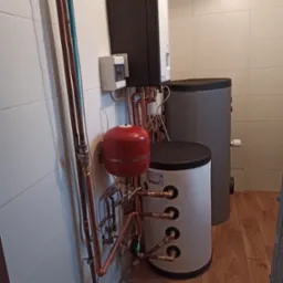 Kompaktowa kotłownia wewnątrz budynku z białymi kafelkami na ścianach, widoczne zbiorniki akumulacyjne, piec, naczynie przeponowe i instalacja z rur miedzianych.